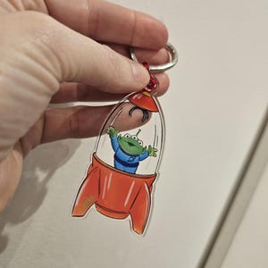 Puede incluir: Un llavero de cohete rojo y naranja con un personaje extraterrestre verde de tres ojos en su interior. El cohete est&aacute; hecho de acr&iacute;lico transparente y tiene un anillo plateado.
