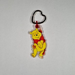 Puede incluir: Un llavero de dibujos animados de Winnie the Pooh de color amarillo con una camisa roja. El llavero tiene un anillo de coraz&oacute;n plateado.
