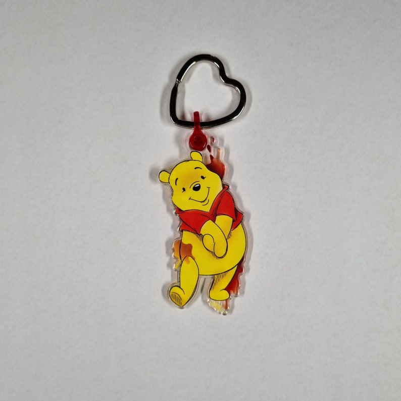 Puede incluir: Un llavero de dibujos animados de Winnie the Pooh de color amarillo con una camisa roja. El llavero tiene un anillo de coraz&oacute;n plateado.