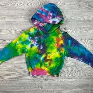 May include: A colorful tie-dye hoodie with a hood and a front pocket. The hoodie features a vibrant mix of colors, including blue, green, yellow, pink, and purple. The garment is laid flat on a wooden surface.