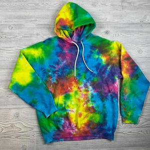 May include: A tie-dye hoodie in a vibrant mix of colors, including yellow, green, blue, red, and purple. The hooded sweatshirt features a front pocket and white drawstrings. The garment is displayed on a light wood-grain surface.