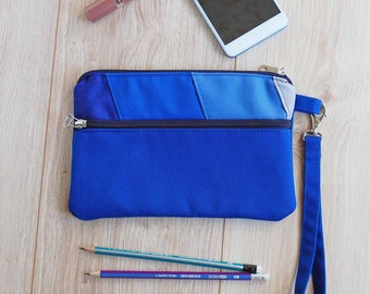 May include: A blue zippered pouch with a wrist strap. The pouch has two compartments and is made of a patchwork of blue fabrics. Two pencils are lying on a wooden surface next to the pouch.