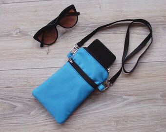 May include: A small blue crossbody bag with a black strap and a zipper closure. A black smartphone is inside the bag. A pair of black sunglasses is lying on a wooden surface.