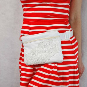 May include: A white leather fanny pack with a white lace design. The fanny pack is worn over a red and white striped dress.