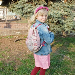 May include: A young girl wearing a pink floral headband, a denim jacket, and a pink skirt with a white floral pattern. She is carrying a drawstring bag with the text "Made in Italy" printed on it.