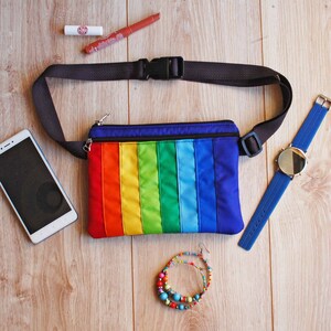 May include: A rainbow-colored fanny pack with a black adjustable strap. The fanny pack is lying on a wooden surface with a cell phone, lipstick, a watch, and a beaded bracelet.