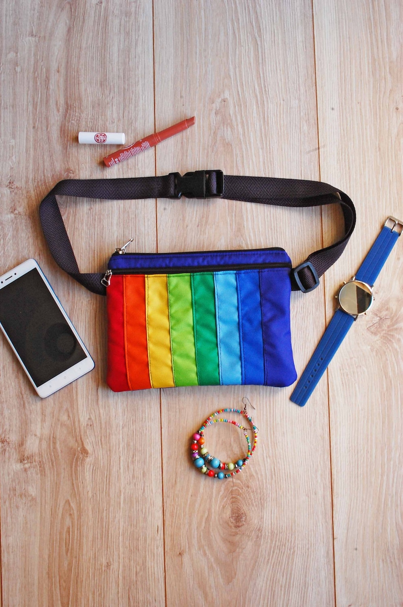 May include: A rainbow-colored fanny pack with a black adjustable strap. The fanny pack is lying on a wooden surface with a cell phone, lipstick, a watch, and a beaded bracelet.