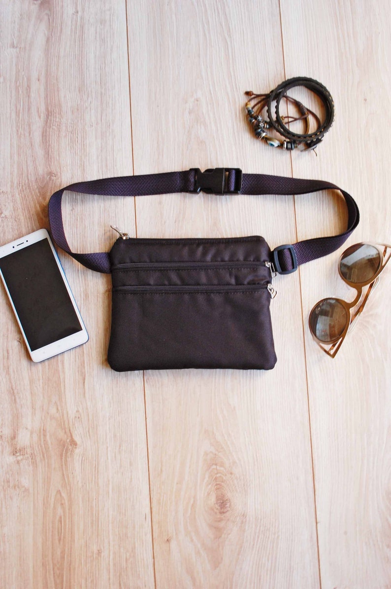 May include: A black fanny pack with a zippered closure and adjustable strap. The fanny pack is laying on a wooden surface with a cell phone, sunglasses, and bracelets.