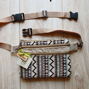 May include: A beige fanny pack with a geometric pattern in black, white, pink, and brown. The pack has three zippered compartments and a brown adjustable strap with black buckles. A small wooden tag with the text "HAND MADE" and a white tag with the text "Fairy Beaches" are attached to the pack.