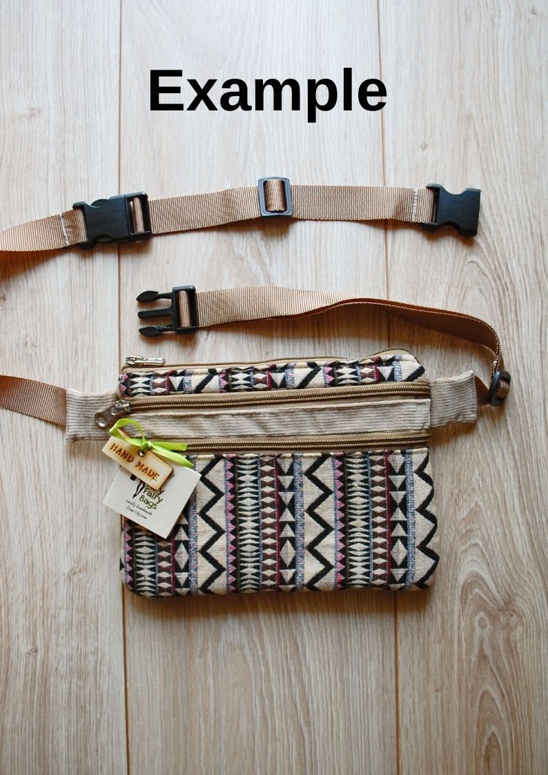 May include: A beige fanny pack with a geometric pattern in black, white, pink, and brown. The pack has three zippered compartments and a brown adjustable strap with black buckles. A small wooden tag with the text "HAND MADE" and a white tag with the text "Fairy Beaches" are attached to the pack.