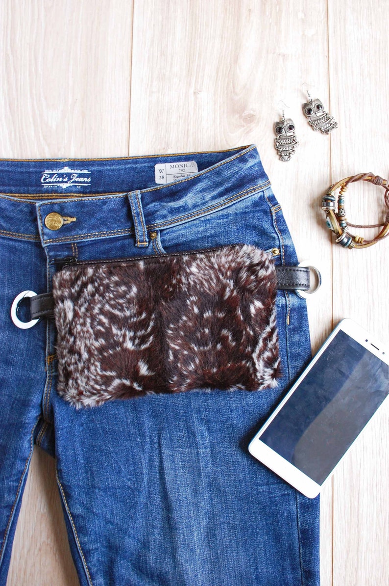 K&ouml;nnte beinhalten: Eine braune und wei&szlig;e Pelz-H&uuml;fttasche mit einem schwarzen Riemen, der an einer blauen Jeans befestigt ist. Die Jeans hat ein Etikett mit der Aufschrift "Colin's Jeans" und ein Gr&ouml;&szlig;enetikett mit der Aufschrift "W 28 Monica 792".