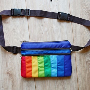 May include: A rainbow-colored fanny pack with a black adjustable strap and two zippered compartments. The pack is made of fabric and has a blue, green, yellow, orange, and red striped design.