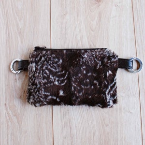 K&ouml;nnte beinhalten: Eine braune und wei&szlig;e Pelz-Bauchtasche mit einem silbernen Rei&szlig;verschluss und silbernen Metallringen an den G&uuml;rtelschlaufen.