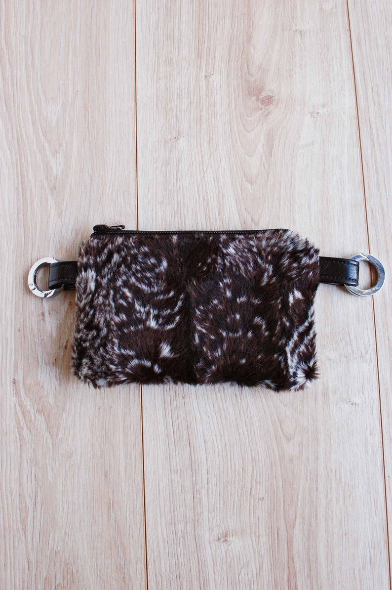 K&ouml;nnte beinhalten: Eine braune und wei&szlig;e Pelz-Bauchtasche mit einem silbernen Rei&szlig;verschluss und silbernen Metallringen an den G&uuml;rtelschlaufen.
