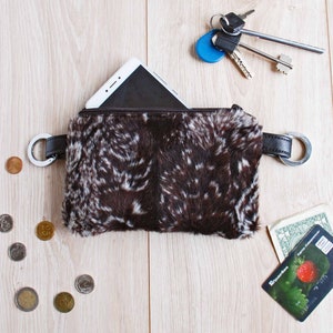 K&ouml;nnte beinhalten: Eine braune und wei&szlig;e Kunstfelltasche mit Rei&szlig;verschluss und einem Metallring an jeder Seite. Ein Smartphone befindet sich in der Tasche. Auf einer Holzoberfl&auml;che liegen Schl&uuml;ssel und eine Kreditkarte.