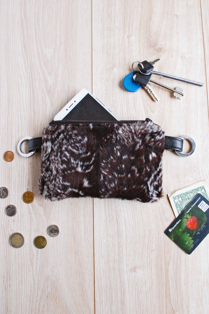 K&ouml;nnte beinhalten: Eine braune und wei&szlig;e Kunstfelltasche mit Rei&szlig;verschluss und einem Metallring an jeder Seite. Ein Smartphone befindet sich in der Tasche. Auf einer Holzoberfl&auml;che liegen Schl&uuml;ssel und eine Kreditkarte.