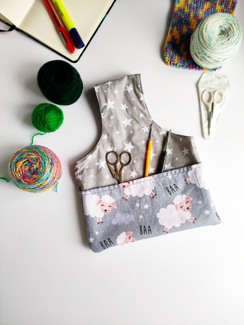 Pode incluir: Uma bolsa de projeto de tecido cinza com um padr&atilde;o de estrelas brancas e uma estampa de ovelhas. A bolsa tem dois bolsos e inclui um par de tesouras, um gancho de croch&ecirc; e uma agulha de tric&ocirc;. A bolsa tamb&eacute;m tem a palavra "BAA" impressa.