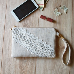 May include: A beige clutch bag with a white lace trim and a gold chain strap. The bag is on a wooden surface with a mobile phone, lipstick, and earrings.