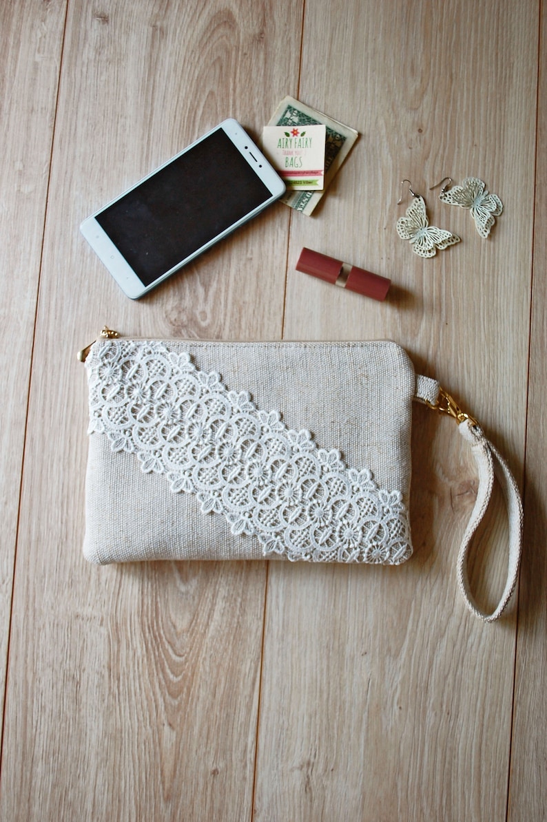 May include: A beige clutch bag with a white lace trim and a gold chain strap. The bag is on a wooden surface with a mobile phone, lipstick, and earrings.