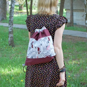 May include: A woman wearing a brown and black floral dress with a drawstring bag featuring a white and red design with houses and cats. The bag has the text "Un Song" repeated on it.
