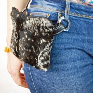 K&ouml;nnte beinhalten: Eine schwarze und wei&szlig;e Pelz-Bauchtasche mit einem schwarzen Riemen und einer silbernen Schnalle. Die Bauchtasche wird um die Taille einer Person getragen, die eine blaue Jeans tr&auml;gt.