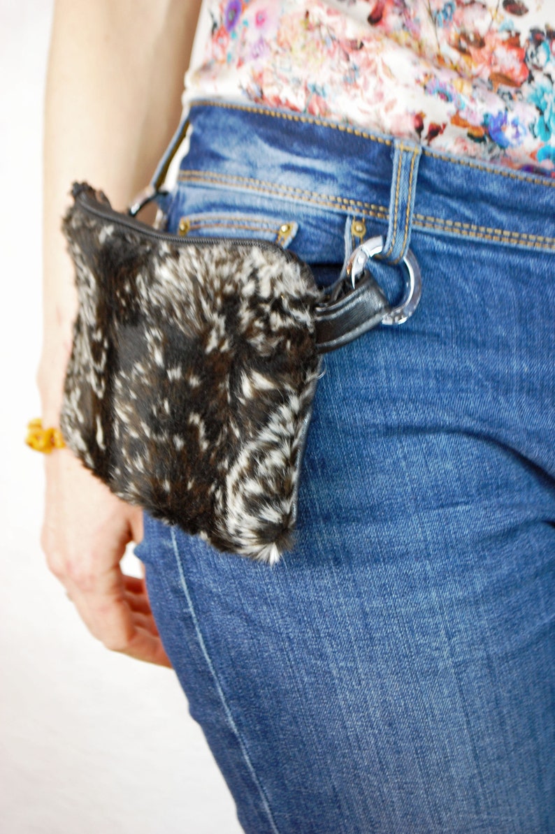 K&ouml;nnte beinhalten: Eine schwarze und wei&szlig;e Pelz-Bauchtasche mit einem schwarzen Riemen und einer silbernen Schnalle. Die Bauchtasche wird um die Taille einer Person getragen, die eine blaue Jeans tr&auml;gt.