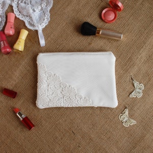 May include: A white makeup bag with a lace trim, two pairs of white butterfly earrings, two red lipsticks, a small bottle of blush, and a makeup brush on a brown burlap background.
