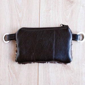 K&ouml;nnte beinhalten: Eine braune Tasche aus Kunstleder mit Rei&szlig;verschluss, pelzigem Besatz und silbernen D-Ringen an den Seiten.