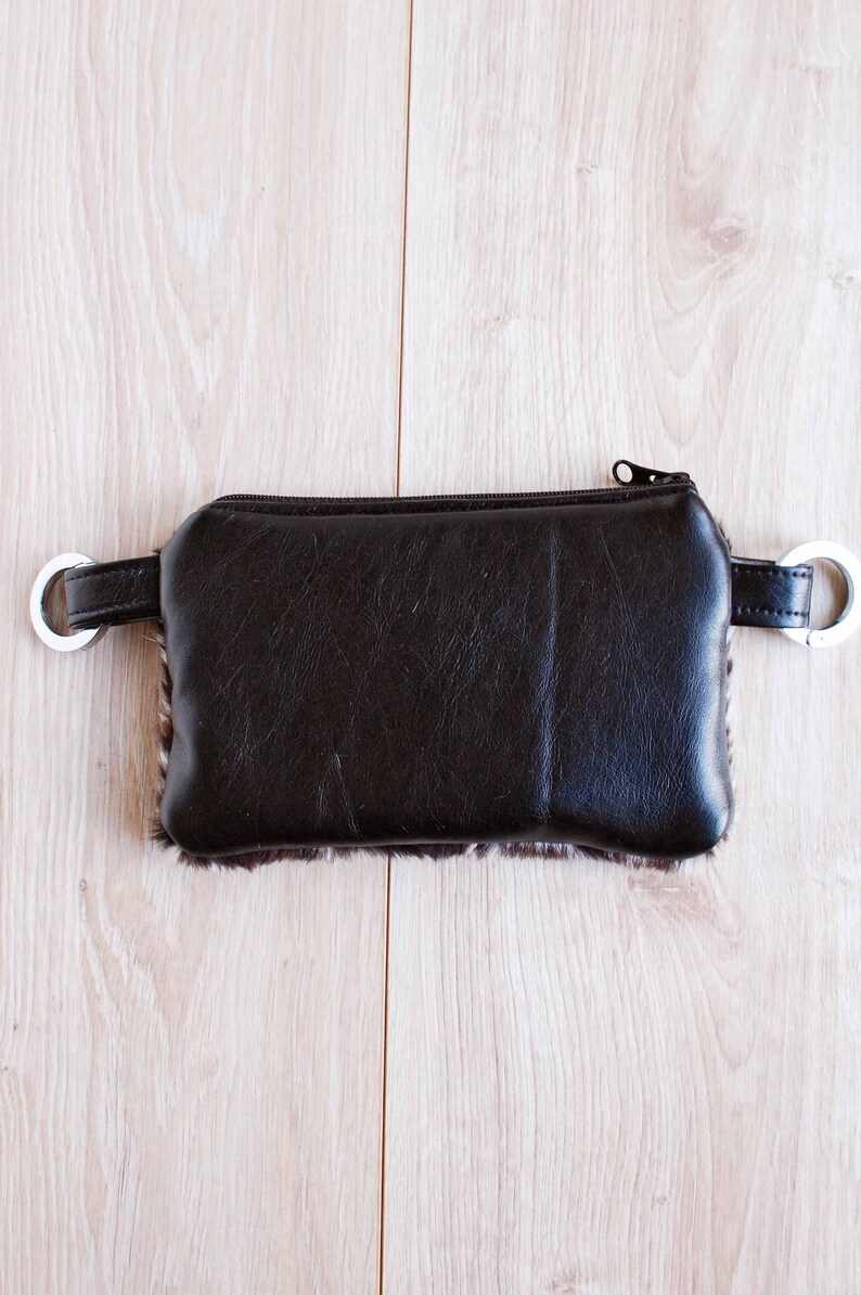 K&ouml;nnte beinhalten: Eine braune Tasche aus Kunstleder mit Rei&szlig;verschluss, pelzigem Besatz und silbernen D-Ringen an den Seiten.