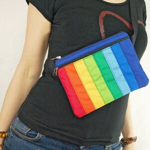 May include: A rainbow striped fanny pack with a black strap worn over a black t-shirt and blue jeans.