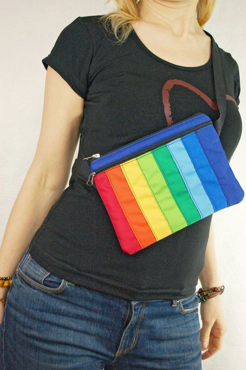 May include: A rainbow striped fanny pack with a black strap worn over a black t-shirt and blue jeans.