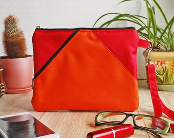 May include: A red and orange zipped pouch with a black zipper and a black strap. The pouch is on a wooden surface with a mobile phone, lipstick, and glasses.