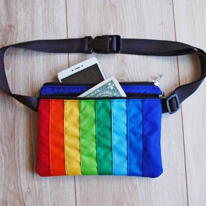 May include: A rainbow-colored fanny pack with a black adjustable strap. The pack has a zippered pocket and is shown with a smartphone and a dollar bill inside.
