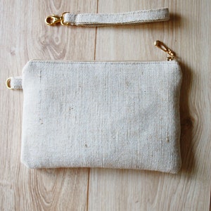May include: A white fabric clutch with a gold zipper and a matching strap. The clutch is made of a textured fabric and has a simple, minimalist design.