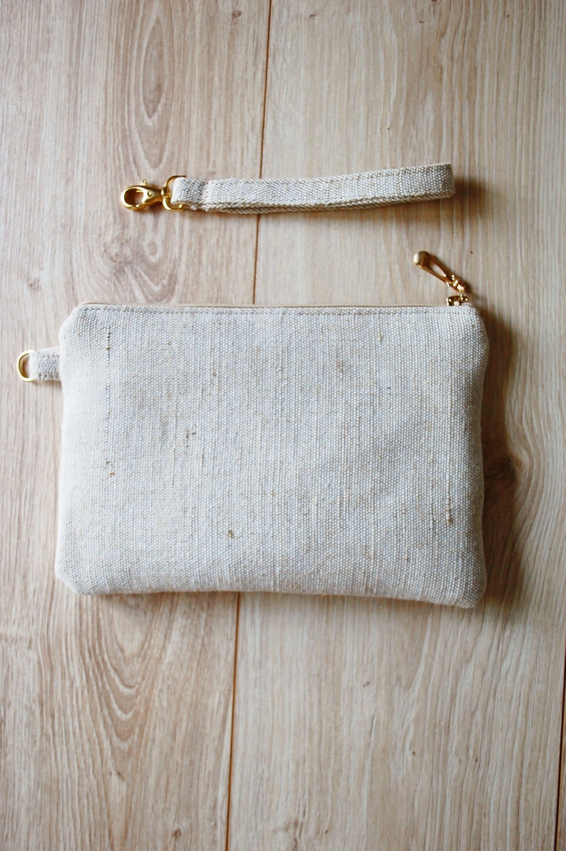 May include: A white fabric clutch with a gold zipper and a matching strap. The clutch is made of a textured fabric and has a simple, minimalist design.