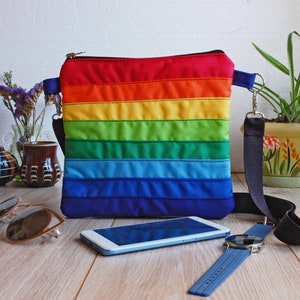 May include: A rainbow striped crossbody bag with a zipper closure and a black adjustable strap. The bag is laying on a wooden surface with a cell phone, sunglasses, and a watch.
