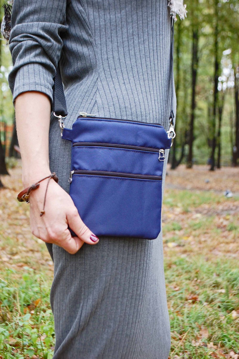 May include: A small blue crossbody bag with three zippered compartments. The bag is worn by a person in a gray ribbed dress.