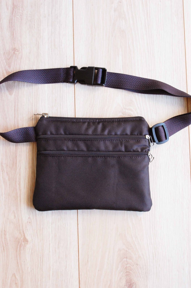 May include: A brown fanny pack with two zippered compartments and a black adjustable strap with a plastic buckle.