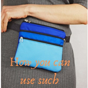 May include: A blue and light blue fanny pack with three zippered compartments. The fanny pack is worn around the waist of a person wearing a gray sweater.