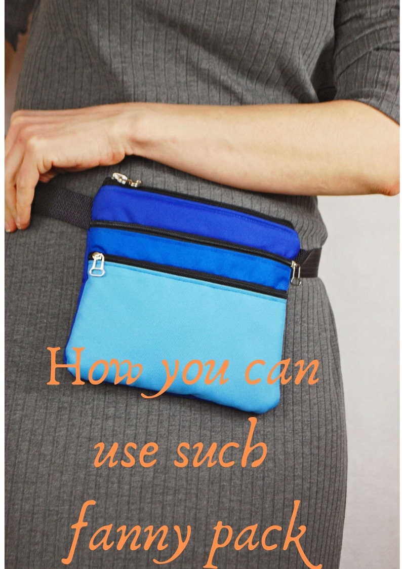 May include: A blue and light blue fanny pack with three zippered compartments. The fanny pack is worn around the waist of a person wearing a gray sweater.