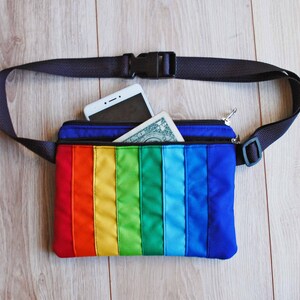 May include: A rainbow-colored fanny pack with a black adjustable strap. The pack has a zippered pocket and is open to reveal a white smartphone and a dollar bill.
