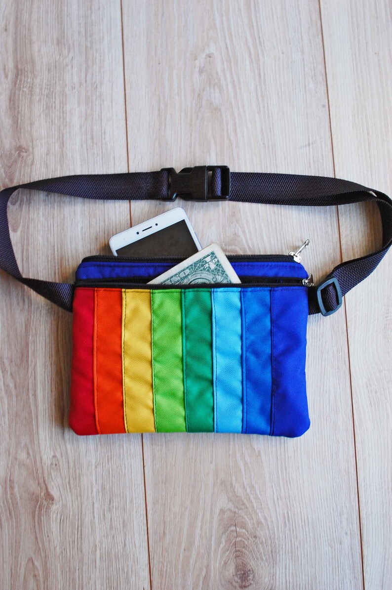 May include: A rainbow-colored fanny pack with a black adjustable strap. The pack has a zippered pocket and is open to reveal a white smartphone and a dollar bill.