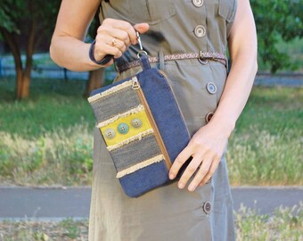 May include: A woman is holding a blue denim clutch with a yellow and brown fabric accent. The clutch has a zipper closure and a brown strap.