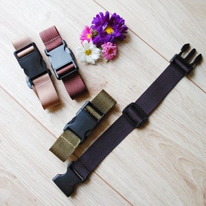 May include: Four different colored straps with black plastic buckles. The straps are brown, beige, green, and black. The straps are lying on a light wood surface.