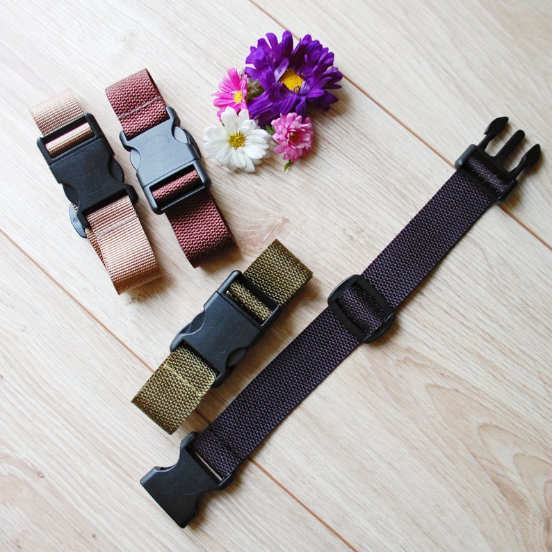 May include: Four different colored straps with black plastic buckles. The straps are brown, beige, green, and black. The straps are lying on a light wood surface.