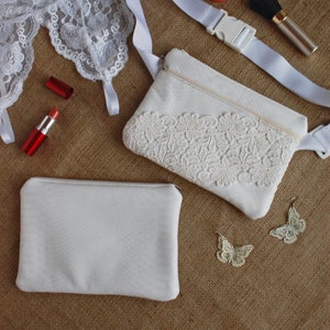May include: Two white fabric pouches with lace trim, one with a zipper and a white strap, and the other with a plain front.  There are two white lace butterflies, two red lipsticks, a red compact, and a makeup brush on a brown hessian background.