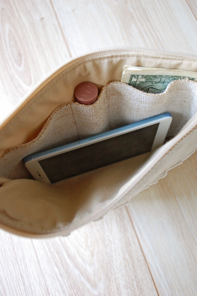 May include: A beige canvas zippered pouch with multiple pockets. The pouch is open and contains a smartphone, a small tube of lip balm, and a one dollar bill.