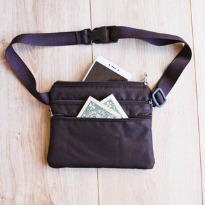 May include: A black fanny pack with a zippered pocket and a white smartphone inside. The fanny pack has a black adjustable strap with a plastic buckle.