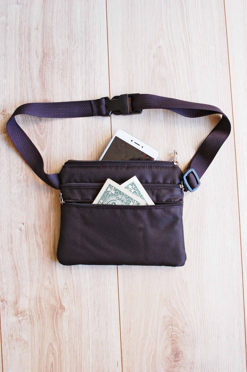 May include: A black fanny pack with a zippered pocket and a white smartphone inside. The fanny pack has a black adjustable strap with a plastic buckle.
