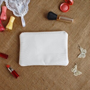 Peut inclure: Une pochette zippée en toile blanche sur un fond en toile de jute brune. Divers articles de maquillage, dont du vernis à ongles, du rouge à lèvres, du fard à joues et un pinceau à maquillage, sont éparpillés autour de la pochette.